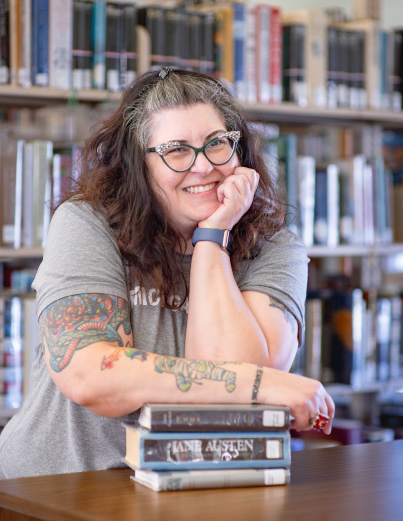 Image of a woman with long hair and smiling. She's leaning on a pile of books.